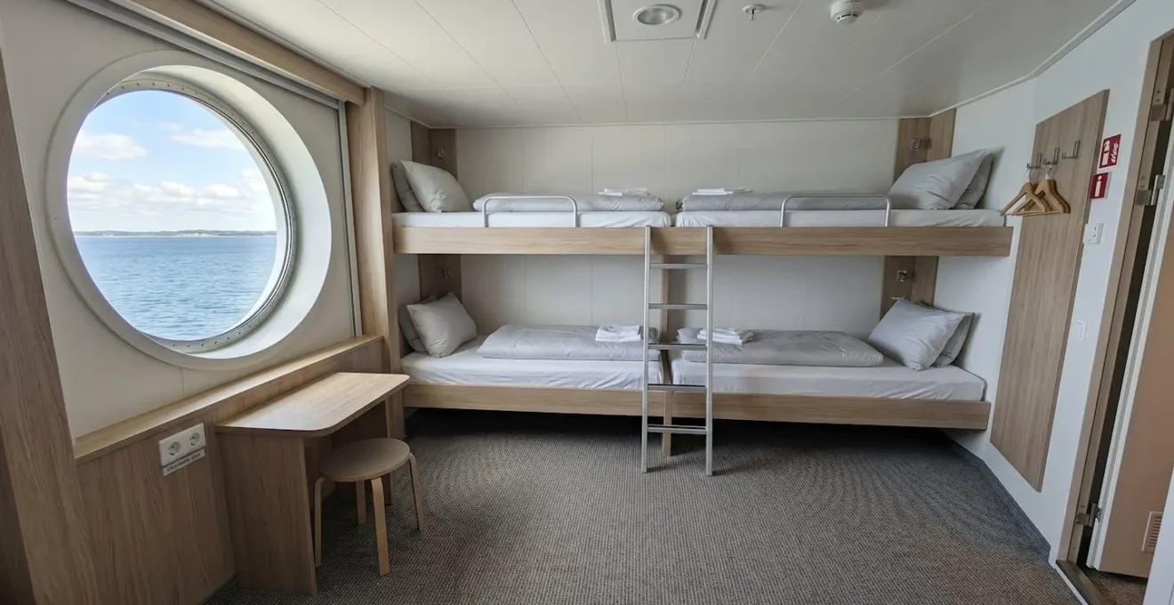 Weitwinkelaufnahme einer leeren, modernen Fährkabine mit vorbereiteten Betten und einem Bullauge, durch das helles Tageslicht einfällt